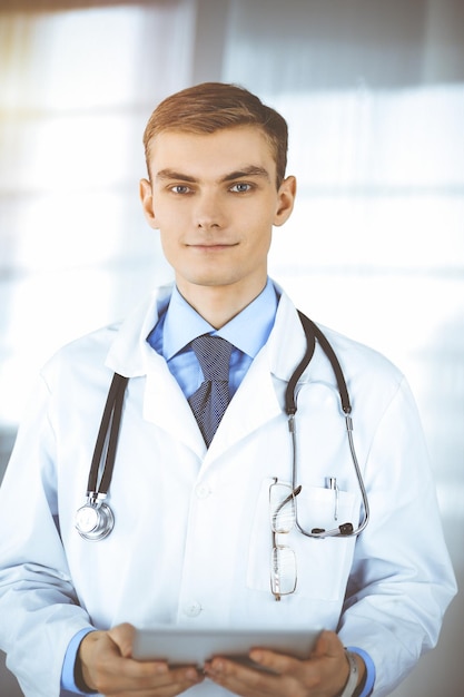 Homem jovem médico está sorrindo alegremente para a câmera, em pé em um escritório de hospital ensolarado, segurando um tablet de computador nas mãos. Serviço médico perfeito na clínica. Futuro feliz em medicina e hea