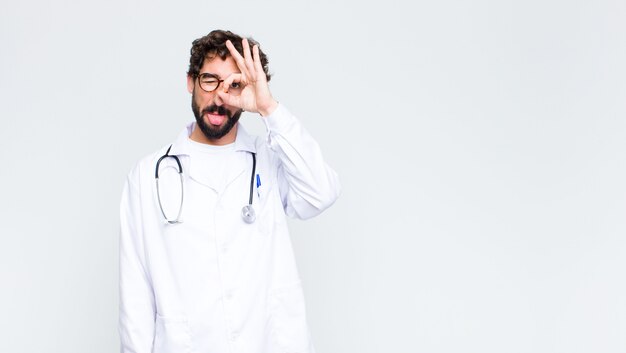 Homem jovem médico sorrindo alegremente com cara engraçada, brincando e olhando pelo olho mágico, espionando segredos contra a parede do espaço de cópia