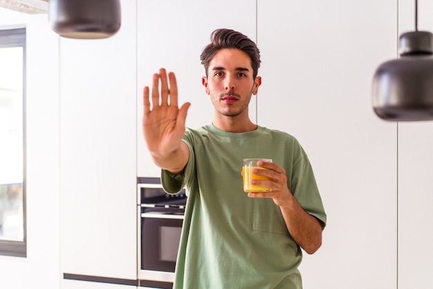 Homem jovem mestiço bebendo suco de laranja em sua cozinha, de pé com a mão estendida, mostrando o sinal de pare, impedindo você.