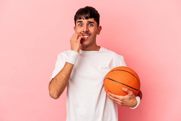 Homem jovem mestiço jogando basquete isolado no fundo rosa, roendo as unhas, nervoso e muito ansioso.