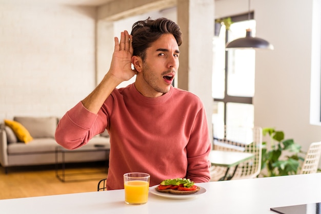 Homem jovem mestiço tomando café da manhã em sua cozinha, tentando ouvir uma fofoca.