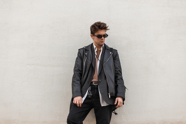 Homem jovem moderno em uma jaqueta de couro superdimensionada elegante em jeans elegantes em óculos de sol escuros com penteado em pé perto de uma parede moderna ao ar livre. Cara atraente hipster em roupas da moda da cidade.