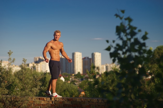 Homem jovem musculoso em pé no topo do telhado e olhando durante o pôr do sol. Conceito de estilo de vida saudável e confiante.