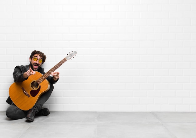 Homem jovem músico, sentindo-se feliz e confiante, apontando para a câmera com as duas mãos e rindo, escolhendo-o com uma guitarra