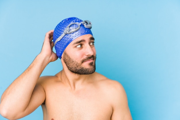 Homem jovem nadador bonito isolado tocando atrás da cabeça, pensando e fazendo uma escolha.