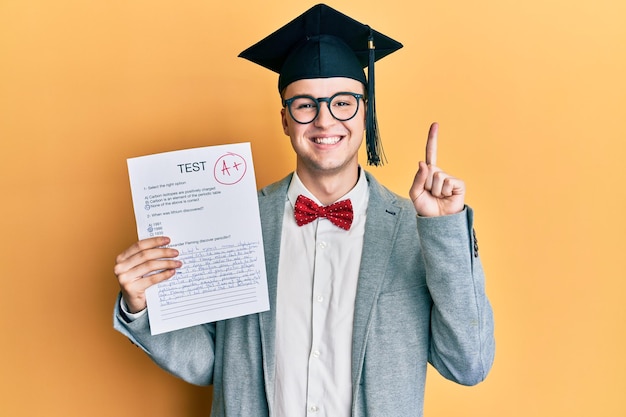 Homem jovem nerd caucasiano usando óculos e boné de formatura segurando exame aprovado sorrindo com uma ideia ou pergunta dedo apontando com cara feliz, número um