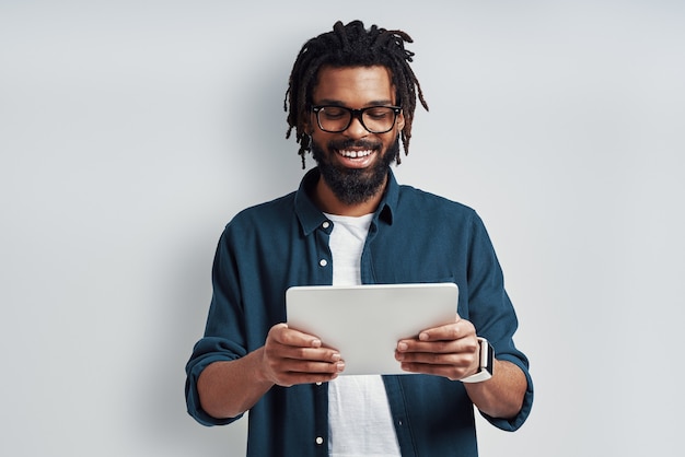 Homem jovem ocupado usando óculos usando tablet digital e sorrindo enquanto fica de pé contra uma parede cinza