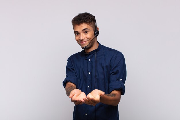 Homem jovem operador de telemarketing sorrindo feliz com um olhar amigável, confiante e positivo, oferecendo e mostrando um objeto ou conceito