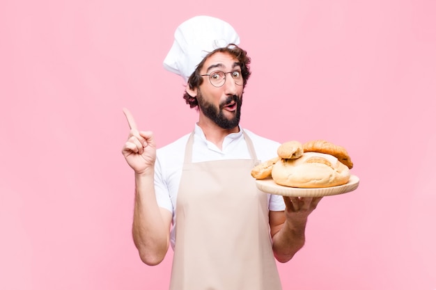 Homem jovem padeiro louco segurando o pão contra a parede rosa