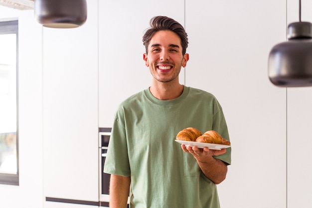 Homem jovem raça mista comendo croissant em uma cozinha pela manhã, rindo e se divertindo.