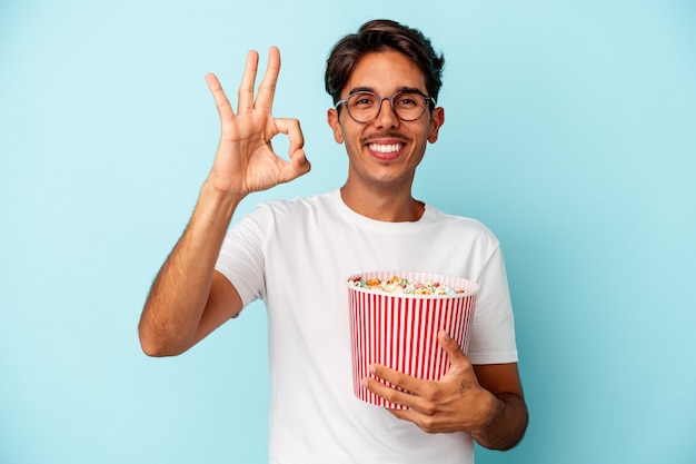 Homem jovem raça mista comendo pipocas isoladas em fundo azul alegre e confiante, mostrando o gesto ok.