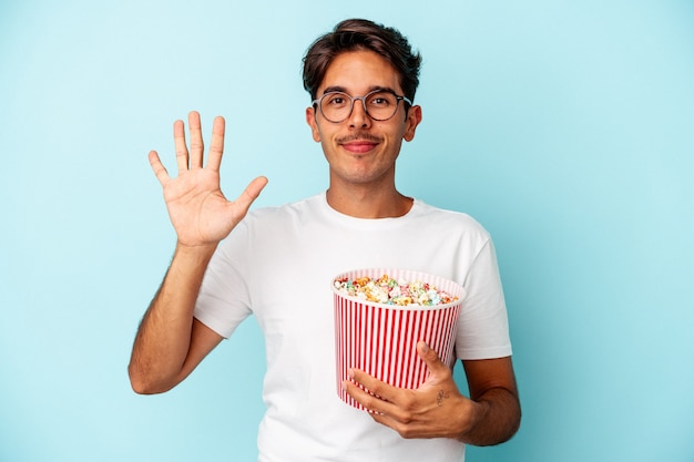 Homem jovem raça mista comendo pipocas isoladas em fundo azul, sorrindo alegre mostrando o número cinco com os dedos.