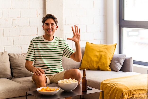 Homem jovem raça mista comendo pipocas, sentado no sofá, sorrindo alegre mostrando o número cinco com os dedos.