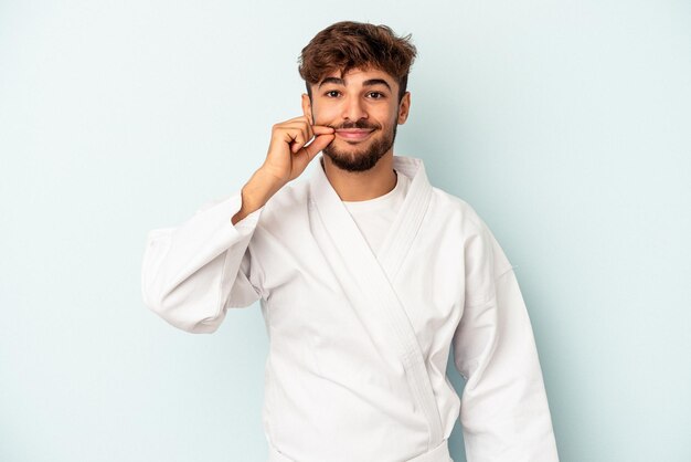 Homem jovem raça mista fazendo caratê isolado em um fundo azul com os dedos nos lábios, mantendo um segredo.