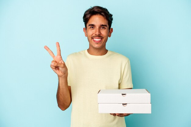 Homem jovem raça mista segurando pizzas isoladas em um fundo azul, mostrando o número dois com os dedos.