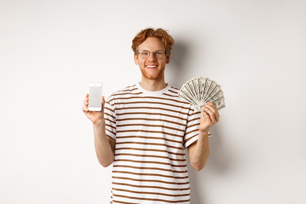 Homem jovem ruiva sorridente de óculos, mostrando a tela em branco do smartphone e dinheiro, em pé sobre um fundo branco.