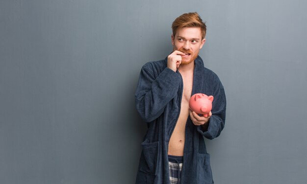 Homem jovem ruiva vestindo pijama relaxado pensando em algo olhando para um espaço de cópia. Ele está segurando um cofrinho.