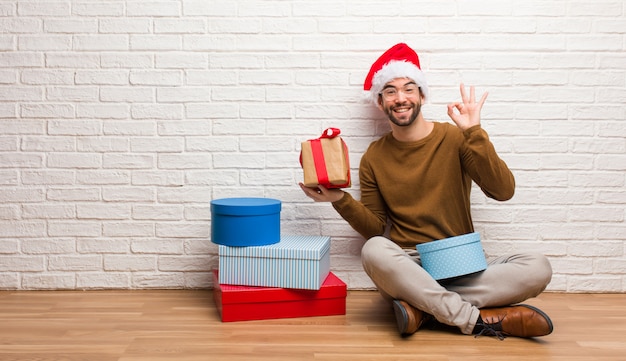 Homem jovem, sentando, com, presentes, celebrando, natal, alegre, e, confiante, fazendo, ok, gesto