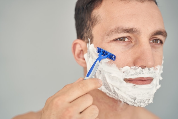 Homem jovem sério e atencioso fazendo a barba com espuma e usando uma navalha descartável