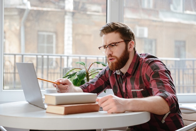 Homem jovem sério perto de livros usando o computador portátil.