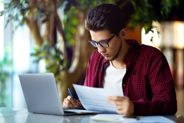 Homem jovem sorridente hipster em copos com o laptop em cima da mesa. programador, desenvolvedor web, designer que trabalha no escritório comparando versões de sites para dispositivos móveis e computadores.