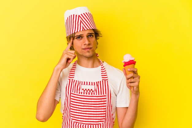 Homem jovem sorveteiro com maquiagem segurando o sorvete isolado no fundo amarelo, apontando o templo com o dedo, pensando, focado em uma tarefa.