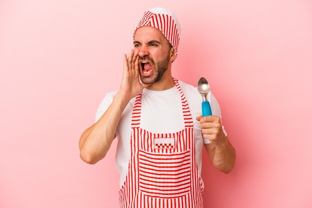 Homem jovem sorveteiro segurando a colher isolada no fundo rosa gritando e segurando a palma da mão perto da boca aberta.