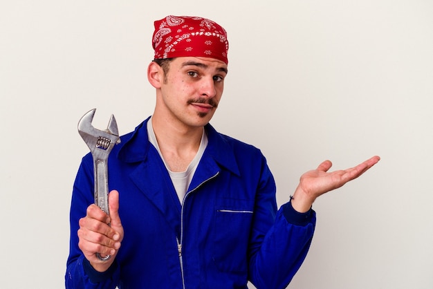 Foto homem jovem trabalhador caucasiano segurando uma chave inglesa isolada no fundo branco, mostrando um espaço de cópia na palma da mão e segurando a outra mão na cintura.