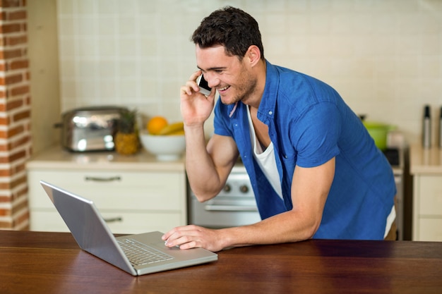 Homem jovem, usando computador portátil, e, conversa telefone, em, cozinha