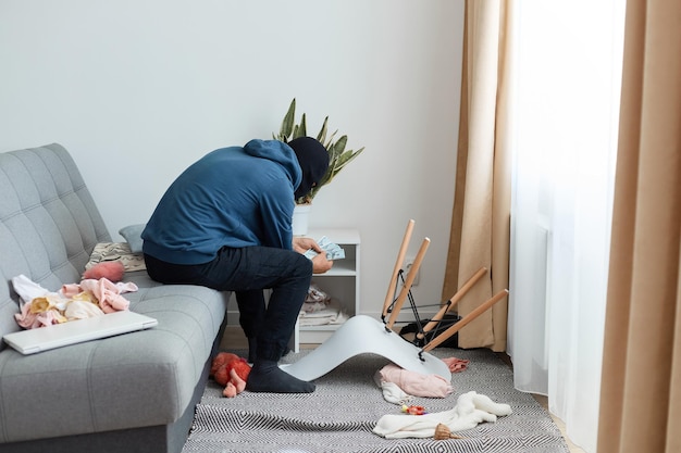 Foto homem ladrão de moletom azul e gorro preto, o ladrão encontrou dinheiro na mesinha de cabeceira da casa que tentando roubar, senta-se no sofá e conta o dinheiro que tem nas mãos.