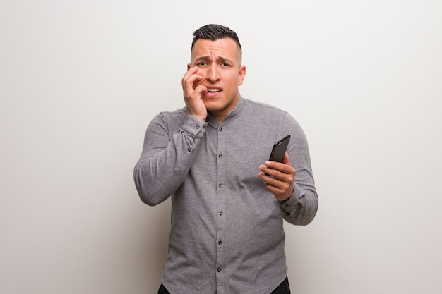 Foto homem latin novo que prende um telefone que morde pregos, nervoso e muito ansioso