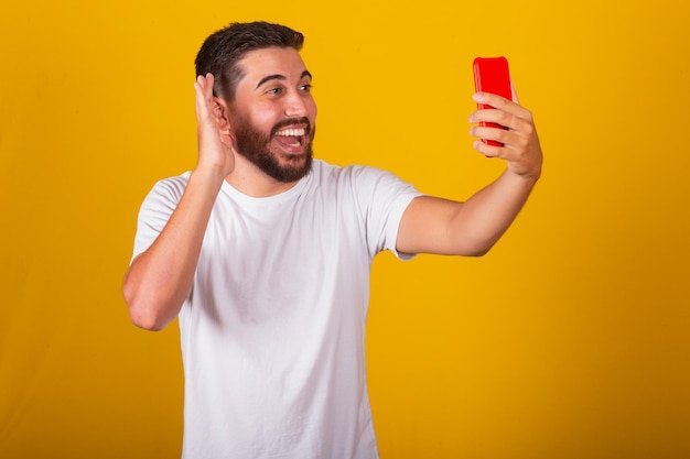 Homem latino-americano brasileiro tomando selfie com celular em videochamada falando sorrindo fotografando fundo amarelo