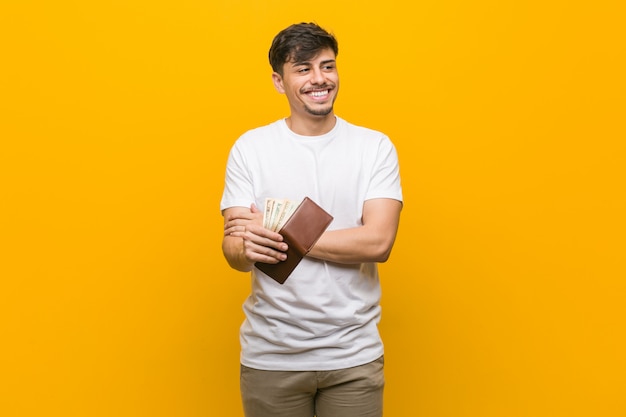 Homem latino-americano novo que mantém um sorriso da carteira seguro com braços cruzados.