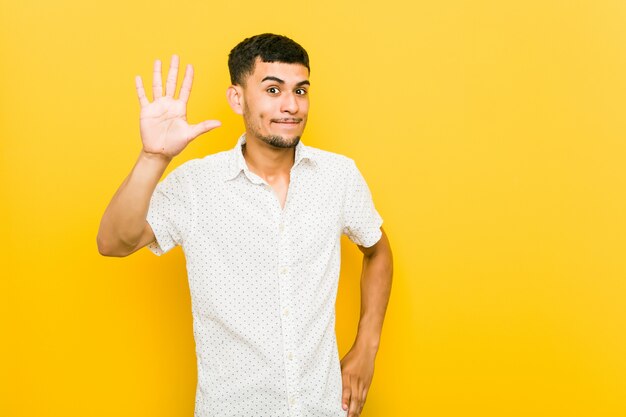 Homem latino-americano novo que sorri mostrando alegre número cinco com dedos.