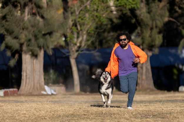 Homem latino com barba, cabelo encaracolado, óculos de sol e roupas casuais correndo com seu cão husky em um parque