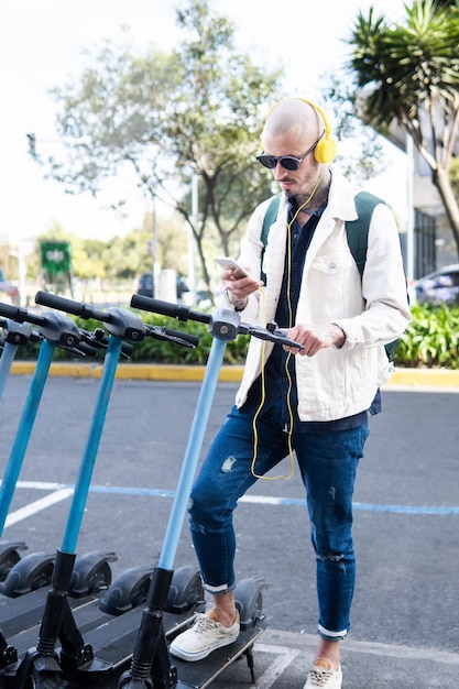 Homem latino com fones de ouvido amarelos e mochila levando uma scooter na cidade com seu celular