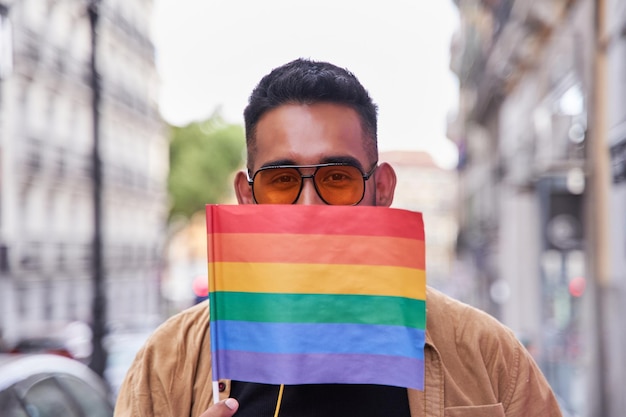 Homem latino com óculos escuros cobrindo a boca com uma bandeira lgbt