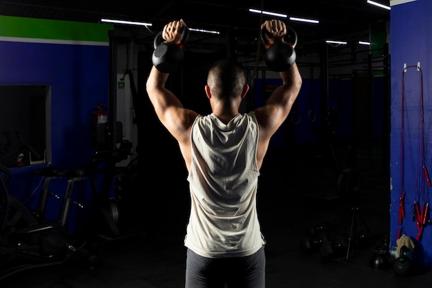 Homem latino com roupas esportivas fazendo imprensa com um par de kettlebells em uma academia