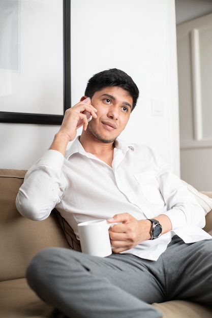 Homem latino de camisa branca no sofá segurando a caneca enquanto fala no smartphone