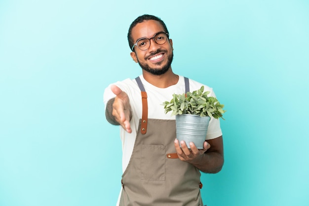 Homem latino de jardineiro segurando uma planta isolada em fundo azul apertando as mãos para fechar um bom negócio