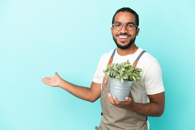 Homem latino de jardineiro segurando uma planta isolada em fundo azul, estendendo as mãos para o lado para convidar para vir