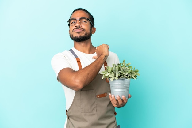 Homem latino de jardineiro segurando uma planta isolada em fundo azul orgulhoso e satisfeito