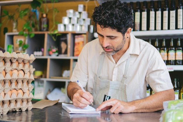 Homem latino de meia-idade fazendo inventário em sua loja orgânica escrevendo em um papel
