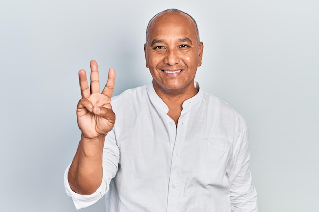 Homem latino de meia-idade vestindo roupas casuais mostrando e apontando para cima com os dedos número três enquanto sorria confiante e feliz