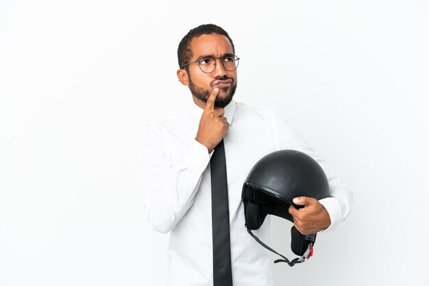 Homem latino de negócios jovem com um capacete de moto isolado no fundo branco com dúvidas ao olhar para cima