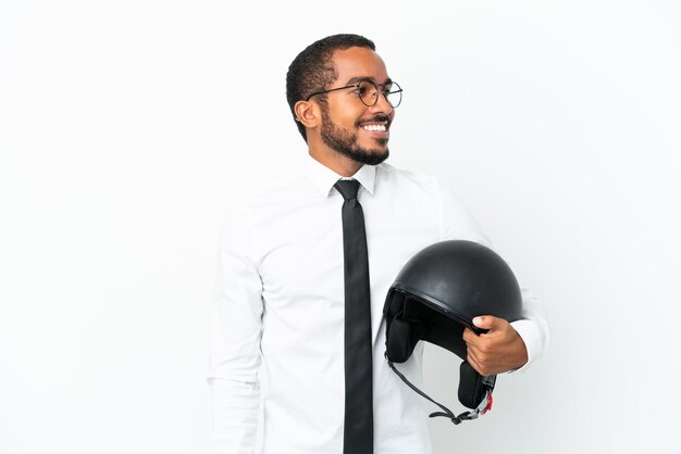 Homem latino de negócios jovem com um capacete de moto isolado no fundo branco, olhando para o lado e sorrindo