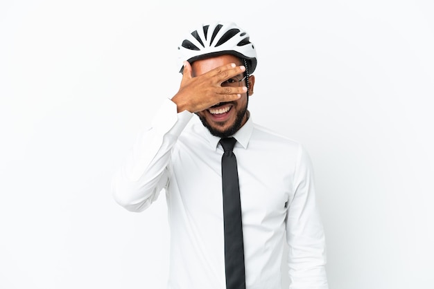 Homem latino de negócios jovem segurando um capacete de bicicleta isolado no fundo branco, cobrindo os olhos pelas mãos e sorrindo