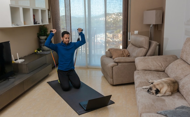 Homem latino fazendo exercícios em sua sala de estar com um elástico enquanto assiste a uma aula online