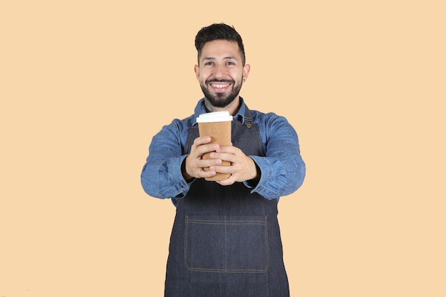 Homem latino-hispânico com avental sorrindo segurando um café com as duas mãos sobre fundo amarelo