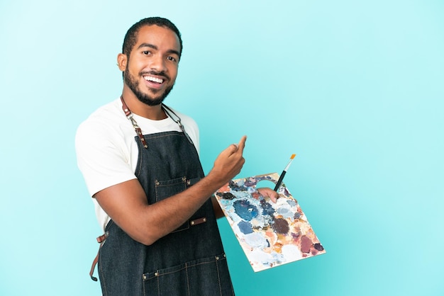 Foto homem latino jovem artista segurando uma paleta isolada em fundo azul, apontando para trás